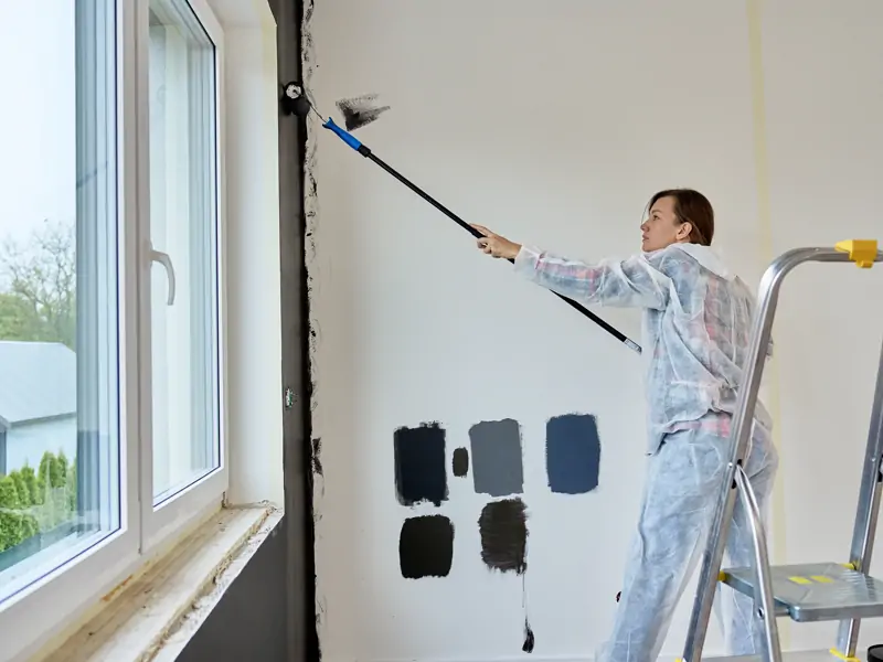 Pintora aplicando tinta na parede com rolo de extensão, ao lado da janela